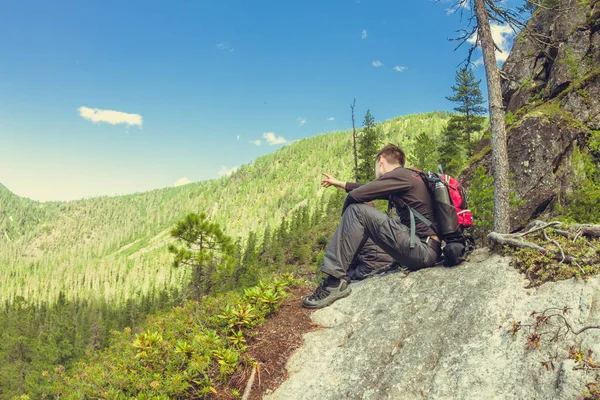 Joven con una mochila se sienta en una roca en la vista de las montañas — Foto de Stock