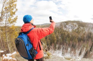 Sırt çantası ve telefonu olan bir yürüyüşçü ormanın üstündeki dağın tepesinde selfie çeker ve vadiye bakar.