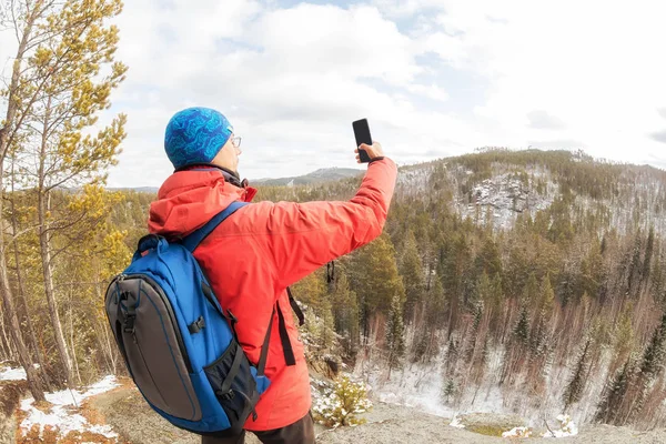 Túrázó férfi hátizsákkal és telefonnal csinál egy szelfit egy hegy tetején az erdő felett, és megnézi a völgyet. — Stock Fotó