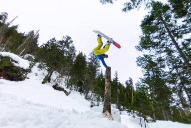 Snowboardcu Freerider, ormanların ve dağların arka planındaki bir kar rampasından atlarken kuru bir kütüğün üzerinde duruyor.