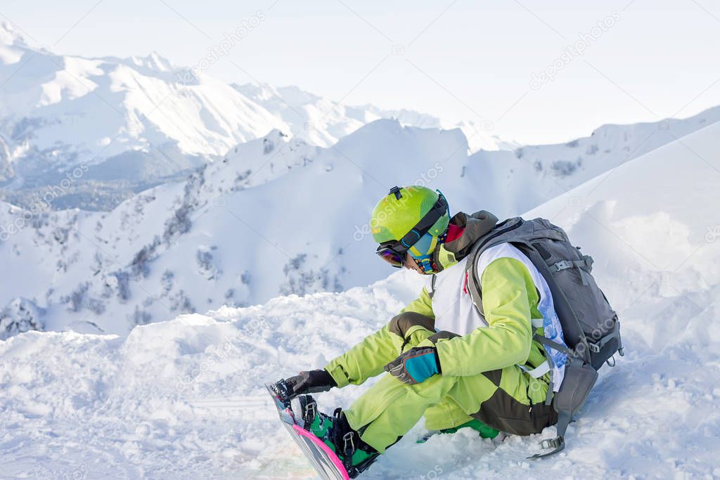 snowboarder freerider sits on a slope and fastens his snowboard while looking at the mountains. Russia Sochi Rosa Khutor