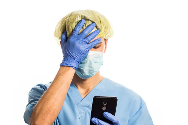 Doctor en ropa médica con guantes de goma y una máscara puso su mano en su cara mientras miraba el teléfono, retrato sobre un fondo blanco — Foto de Stock