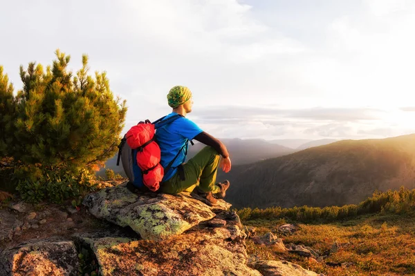 Wanderer mit Rucksack sitzt auf einem Felsen an einem Baum vor dem Hintergrund der Morgendämmerung — Stockfoto