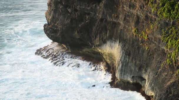 Golven slaan van stenen op een tropisch strand, die een plons vormen. Krachtige golven op een rotsachtige strand. Beroemde Uluwatu tempel in Bali, Indonesië. — Stockvideo