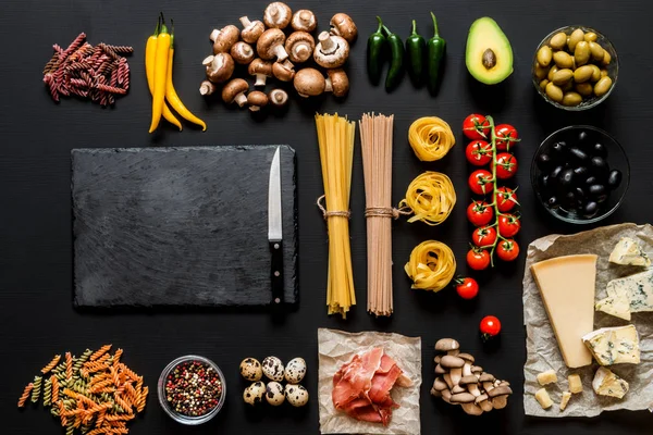 Verschillende verse ingrediënten voor het koken Italiaanse pasta, spaghetti, fettuccine, fusilli en groenten op een zwarte achtergrond met schoolbord en presentexemplaar ruimte. Plat lag, top uitzicht. — Stockfoto