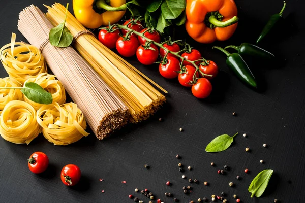 Ruwe Italiaanse pasta. Spaghetti en fettuccine met tomaten, groenten, kruiden en peper op een zwarte achtergrond. Closeup. — Stockfoto