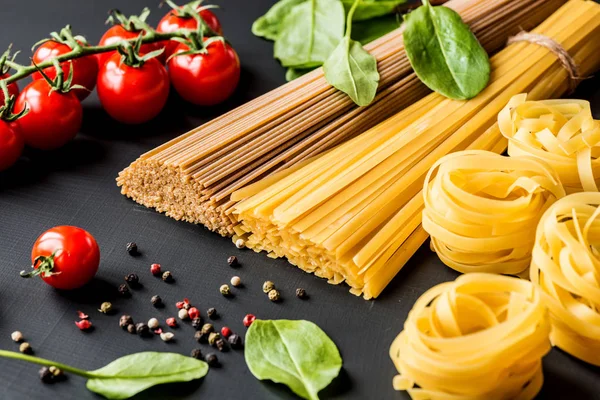 Ruwe Italiaanse pasta. Spaghetti en fettuccine met tomaten, kruiden en peper op een zwarte achtergrond. Closeup. — Stockfoto