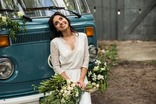Cheerful happy young bride sit on the bumper retro-minibus. Close-up.