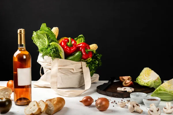 Cotton eco bag with organic different vegetables on a table in the kitchen with a bottle of wine.