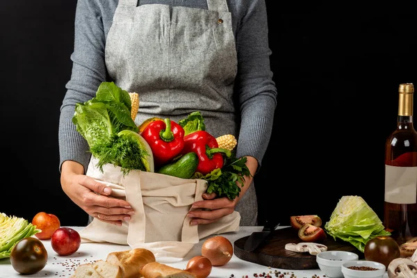 Cotton eco bag with organic different vegetables on a table in the kitchen in the hands of a woman in an apron. Cooking healthy vegetarian food. Zero waste purchases concept.