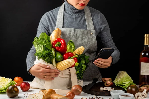 Order food online. Woman in the kitchen holds an eco fabric bag full of fresh organic vegetables and a smartphone in hand. Purchases concept. Cooking healthy food.