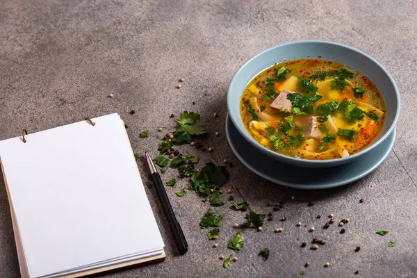 Köstliche würzige Suppe mit Rindfleisch und Nudeln auf grauem Hintergrund. Auf einem Tisch liegt ein Buch zum Schreiben von Rezepten. Nahaufnahme. — Stockfoto