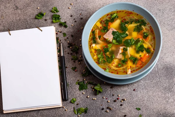 Köstliche würzige Suppe mit Rindfleisch und Nudeln auf grauem Hintergrund. Ein Buch zum Aufschreiben von Rezepten liegt auf dem Tisch. — Stockfoto