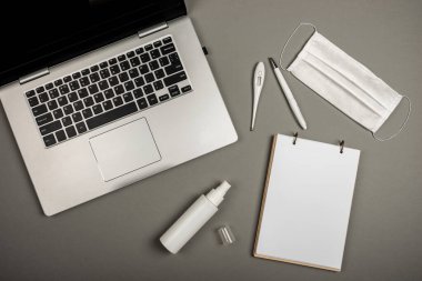 Laptop, defter, kalem, cerrahi maske, tıbbi eldivenler, el dezenfektanı ve gri arka planda dijital termometre. Çevrimiçi iş veya eğitim.