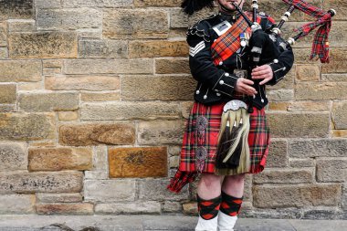 EDINBURGH, SCOTLAND, 24 March 2018 , Scottish bagpiper dressed in traditional red and black tartan dress stand before stone wall. Edinburgh, the most popular tourist city destination in Scotland. clipart