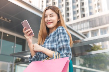 Alışveriş merkezinin arka planında alışveriş torbaları olan gülümseyen Asyalı bir kadın. İnternetten akıllı bir telefon alışverişi yapıyor ve alışveriş merkezi inşaatında gülümsüyor. yaşam tarzı konsepti