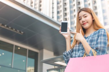 Alışveriş merkezinin arka planında alışveriş torbaları olan gülümseyen Asyalı bir kadın. İnternetten akıllı bir telefon alışverişi yapıyor ve alışveriş merkezi inşaatında gülümsüyor. yaşam tarzı konsepti