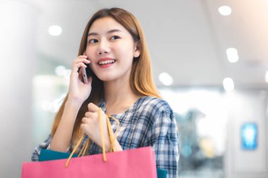 Alışveriş merkezinin arka planında alışveriş torbaları olan gülümseyen Asyalı bir kadın. İnternetten akıllı bir telefon alışverişi yapıyor ve alışveriş merkezi inşaatında gülümsüyor. yaşam tarzı konsepti