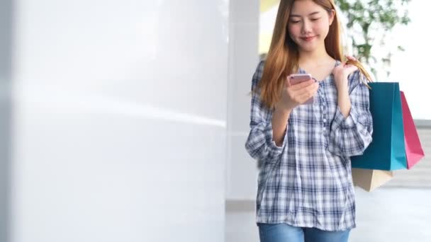 Улыбающаяся Молодая Азиатка Пакетами Покупок Фоне Торгового Центра Используя Смартфон — стоковое видео