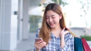 Alışveriş merkezinin arka planında alışveriş torbaları olan gülümseyen Asyalı bir kadın. İnternetten akıllı bir telefon alışverişi yapıyor ve alışveriş merkezi inşaatında gülümsüyor. yaşam tarzı konsepti