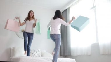Mutlu Asyalı iki genç kadın yatak odasında elinde renkli alışveriş torbalarıyla yatakta zıplarken gülümsüyor. Alışveriş için mutlu ve heyecanlı. Ağır çekim. Mutlu alışveriş zamanı..