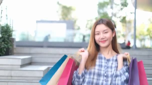 モールの背景にショッピングカラーのバッグで若いアジアの女性を笑顔 スマートフォンのショッピングをオンラインで利用しモールビルに立っていると笑顔になります ライフスタイルのコンセプト — ストック動画