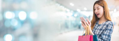 Alışveriş merkezinin arka planında alışveriş torbaları olan gülümseyen Asyalı bir kadın. İnternetten akıllı bir telefon alışverişi yapıyor ve alışveriş merkezi inşaatında gülümsüyor. yaşam tarzı konsepti