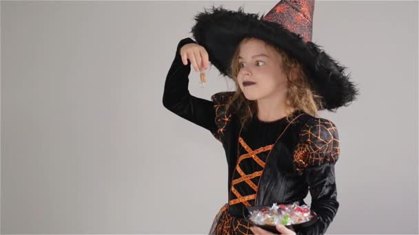 Flicka klädd i en svart-orange klänning och en stor hatt. Flickan har godis i händerna. Trick or Treat, Halloweenfest — Stockvideo