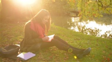 Çimenlerin üzerinde oturan ve bir defter ile güzel park ve göl içinde belgili tanımlık geçmiş yazıları güzel kız