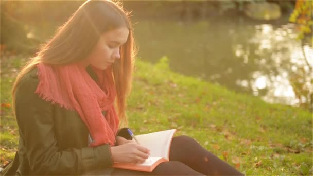 Krásná dívka sedící v trávě a psaní v poznámkovém bloku s krásným parkem a jezerem v pozadí — Stock video
