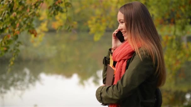 Young female talking on cellphone in the autumn garden near lake, beautiful girl laughing, public park background — Stock Video