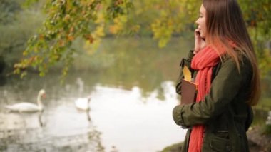 Güzel öğrenci kız cep telefonu üzerinde sonbaharda konuşurken. Sonbahar beyaz bir kadın Smartphone konuşmayı park, Beyaz Kuğu Gölü içinde belgili tanımlık geçmiş Yüzme