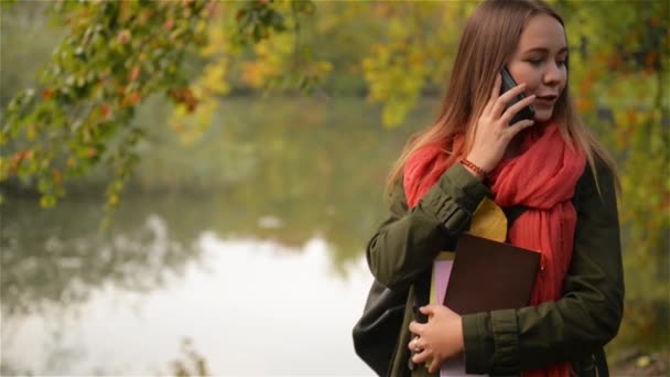 Mooie student meisje praten op mobiele telefoon in de herfst. Herfst Kaukasische vrouw met smartphone gesprek in het park, witte zwanen zwemmen in het meer op de achtergrond — Stockvideo