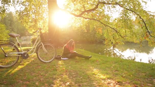 Mooi meisje ontspannen in herfst park leesboek, zittend op het gras in de buurt van lake met haar fiets. Zonnige dag — Stockvideo