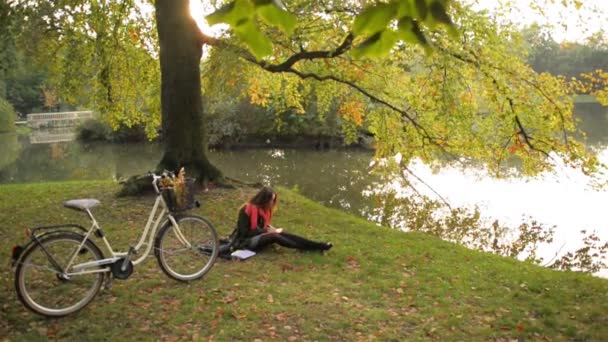 Nöjd student tjej lugnt på grass läser bok i höst offentliga park. Cykel i bakgrunden. — Stockvideo