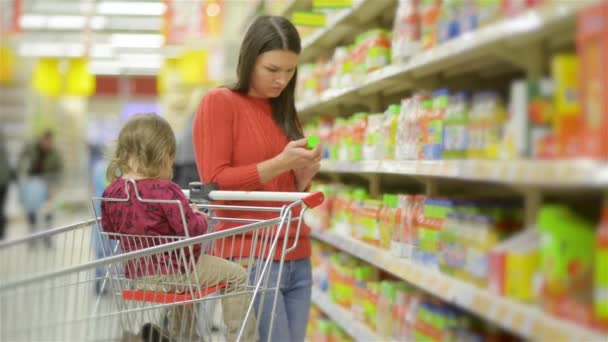 Mutter und Kind gehen an den Großmarktregalen entlang und nehmen Waren im Einkaufswagen, schöne junge Frau kauft im Supermarkt ein — Stockvideo