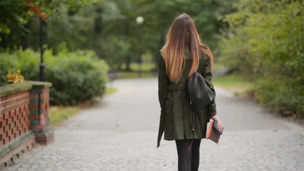 A happy young woman walks on a sidewalk near a city park, student girl holding notebook, warm autumn day — Stockvideo