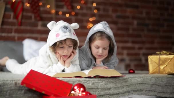 Deux petits frères et sœurs mignons lisant un livre au lit près du sapin de Noël avec des lumières et de l'éclairage. Famille heureuse de deux enfants — Video