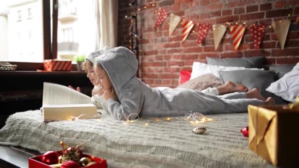 Kinder im warmen Weihnachtsschlafanzug lesen auf dem Bett sitzend ein interessantes Buch, dekorieren Weihnachten und Geschenke