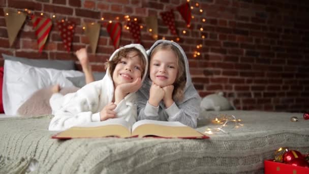Fröhliche süße kleine Kinder sind in warmen Weihnachtsschlafanzug gekleidet lacht und blickt in die Kamera, lächelnde Kinder lesen Buch am Weihnachtsmorgen — Stockvideo