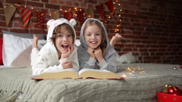 Χαριτωμένο χαμογελώντας αδελφή και ο αδελφός, διαβάζοντας ένα βιβλίο και γελώντας στο σπίτι σε μια εποχή Χριστουγέννων. Βρίσκονται σε αναμονή για ένα Βασίλη. — Αρχείο Βίντεο