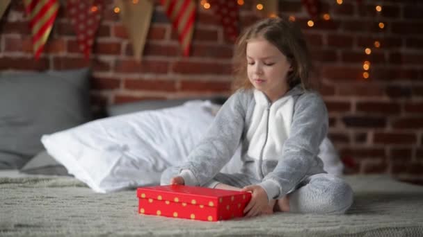 Niña preadolescente despierta en su cama vestida con pijamas de Navidad cálidos en la mañana de vacaciones, disfrutando con regalos, backgraund decorar la Navidad y la iluminación — Vídeos de Stock