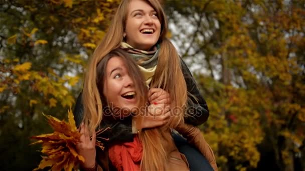 Fröhliche glückliche Frau huckepack ihre hübsche Freundin, zwei schöne junge Mädchen lachen im herbstlichen Park und halten einen Strauß gelber Blätter in der Hand — Stockvideo