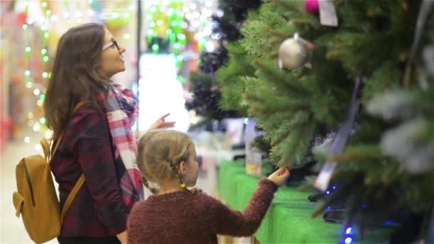スーパーで母と子を選択するクリスマス ツリー。若く美しい母と娘を買うスーパー マーケットの棚、メリー クリスマスと幸せな新年の近くの装飾とクリスマス ツリー — ストック動画