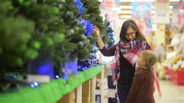 Мать и ребенок выбирают елку в супермаркете. Молодая красивая мама и дочь покупает елку с украшениями возле супермаркета полки, счастливого Рождества и счастливого Нового года — стоковое видео