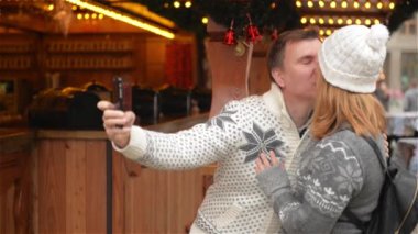 Teknoloji ve insan kavramı. Mutlu çift turist Selfie Smartphone ile Noel piyasada alarak sıcak giysiler içinde. Genç aile gülüyor ve Noel Fuarı öpüşme
