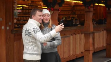 Genç bir çekici çift Selfie Smartphone ile alarak bir Noel pazarda görünümünü. Genç aile Xmas Fuarı gülüyor