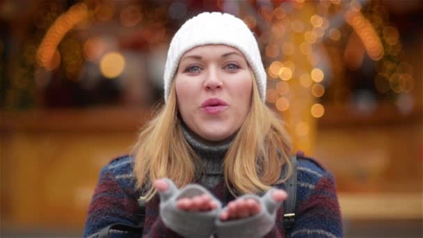 Glückliche unbeschwerte Frau mit blonden Haaren sendet einen Luftkuss über die Weihnachtsbeleuchtung Hintergrund. schönes Mädchen mit weißem Hut und grauen Handschuhen lächelt auf der Neujahrsmesse. — Stockvideo