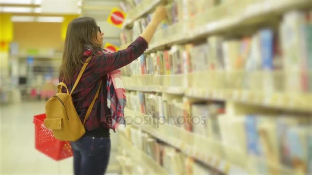 Jeune belle fille portant une écharpe et des lunettes Choisir un livre au supermarché. Femme aux longs cheveux foncés et sac à dos jaune prend un livre et parcourt une page — Video