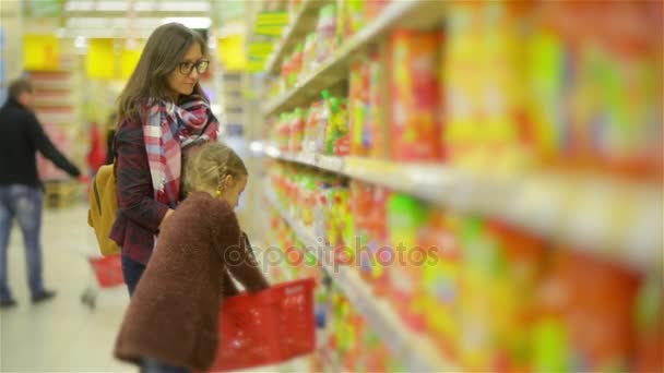 女性と女の子のショッピング モールで商品を選択します。赤いバスケットにいくつかの商品がスーパーの棚に沿って歩くママと配置後の娘 — ストック動画